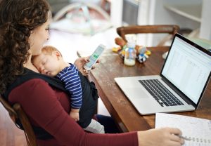 Photo of a woman holding a sleeping infant while checking her cellphone and sitting in front of her laptop
