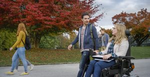 Male student talking with female student sitting on powered wheelchair at university campus.