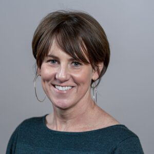 A smiling woman with short brown hair and hoop earrings, wearing a dark green top.