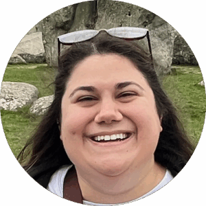 Circular outdoor portrait — laughing woman with shoulder-length dark wavy hair and sunglasses pushed up on her head, in a black jacket over a white top, against a softly blurred stone-and-grass historic site (megalithic stones) background