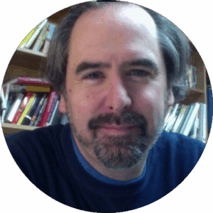 Circular indoor portrait — slightly smiling man with short greying dark hair, a salt-and-pepper goatee, wearing a navy v-neck sweater over a teal t-shirt; bookshelf with multicolored book spines visible behind him