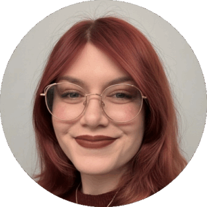 Circular headshot — smiling young woman with shoulder-length bright reddish-auburn hair, round gold wire-frame glasses, dark berry lipstick, wearing a dark red sweater, against a plain light-grey background