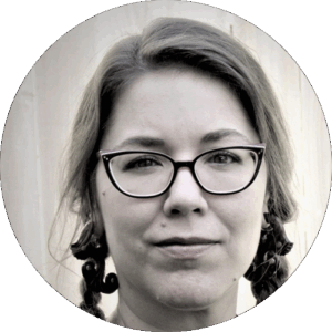 Circular black-and-white headshot — woman with shoulder-length wavy hair partly pulled back into braided sections, dark-framed glasses, and small dangling earrings, faint closed-lip expression, against a neutral light background