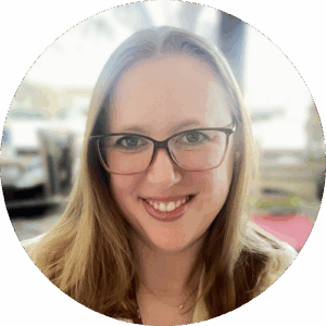 Circular headshot — smiling woman with long blonde hair, wire-frame glasses, and a small pendant necklace, against a softly blurred indoor background
