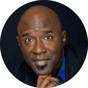 Circular headshot — smiling Black man with shaved head and goatee, resting his chin lightly on his hand, wearing a navy blazer over a bright royal-blue button-up against a black studio background