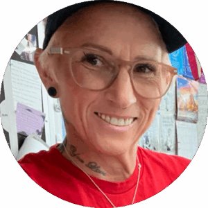Circular outdoor/indoor portrait — smiling person with shaved sides under a black headwrap, tan-framed glasses, black gauge ear plug, and a small chest tattoo visible above a bright red shirt; background shows a wall of sticky notes and small book covers (likely a classroom or library)