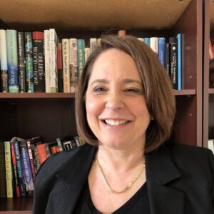 A smiling woman with shoulder-length brown hair, wearing a black blazer, in front of a full bookshelf.
