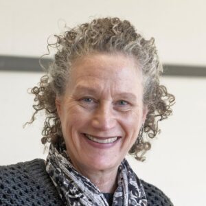 A smiling woman with curly gray hair, wearing a patterned scarf and a dark sweater.